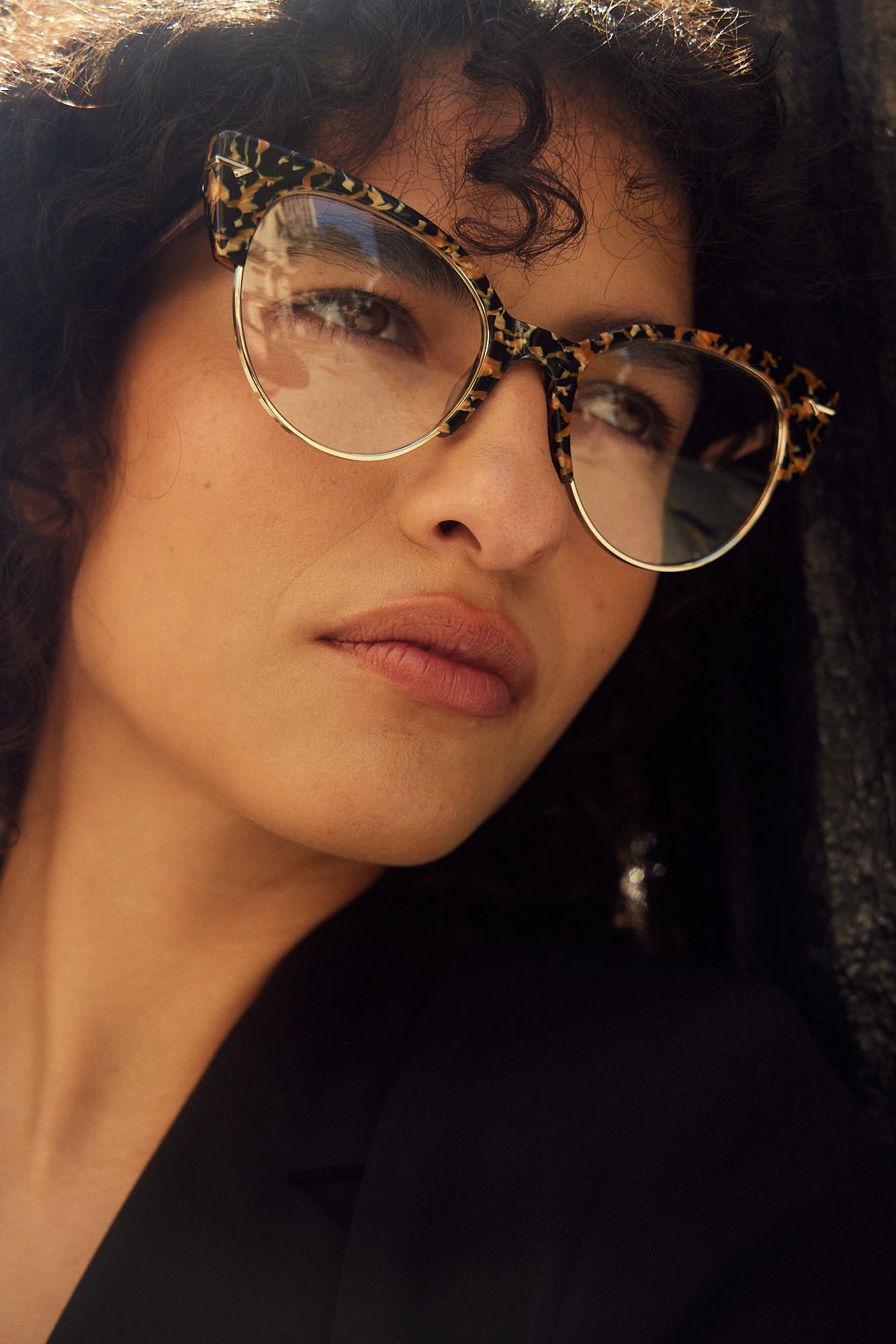 A close-up portrait of a woman with dark curly hair wearing cat-eye eyeglasses with an animal print acetate frame with metal lower rims and rivets.