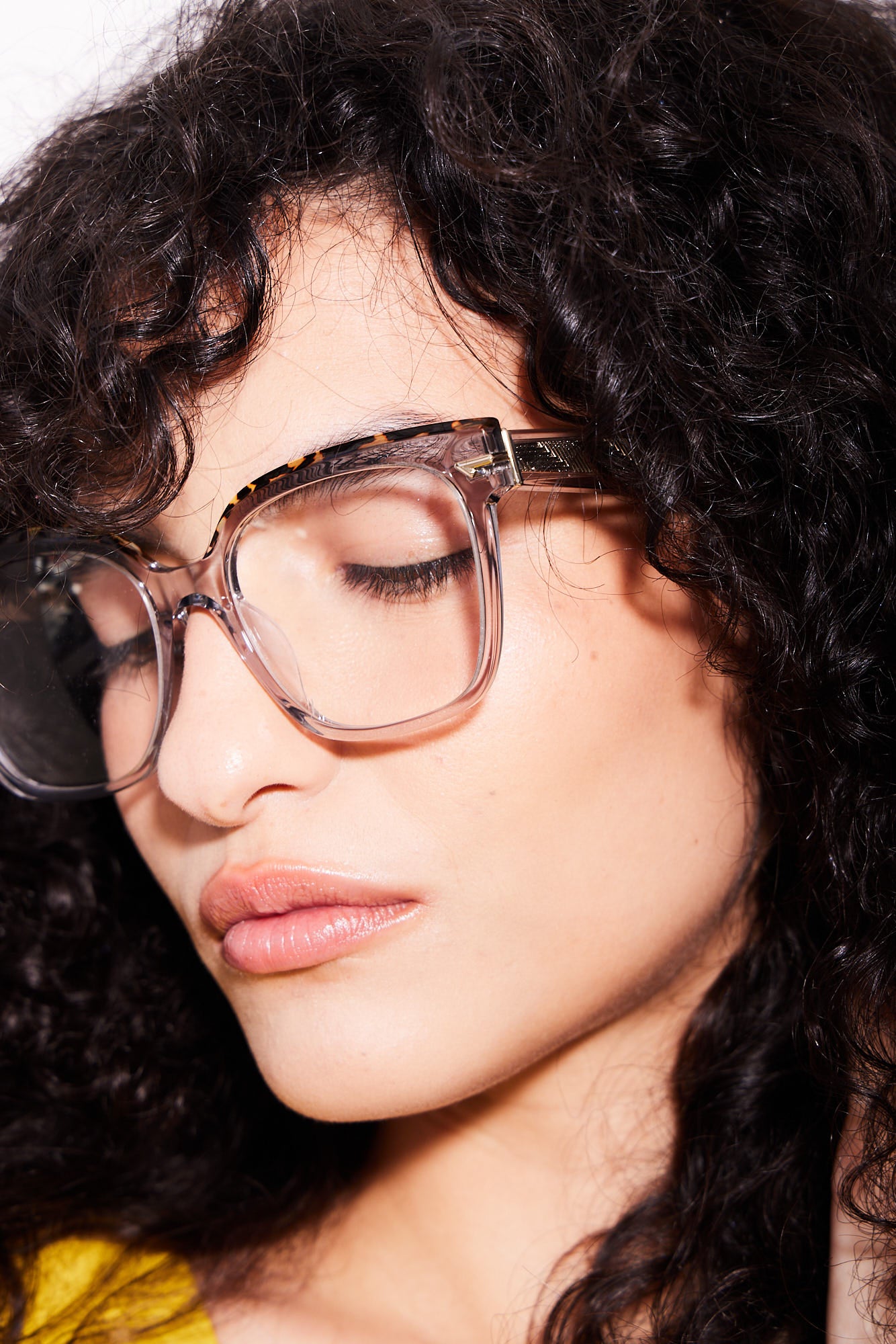 A close-up portrait of a woman with curly dark hair wearing oversized square warm grey acetate eyeglasses with a tortoiseshell top rims.