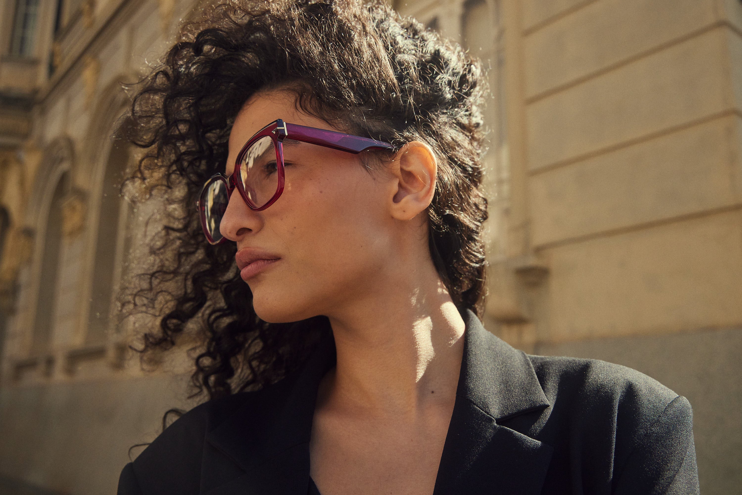 A close-up portrait of a woman with curly dark hair wearing oversized square eyeglasses with purple frames and dark lenses, set against a blurred beige stone building background.