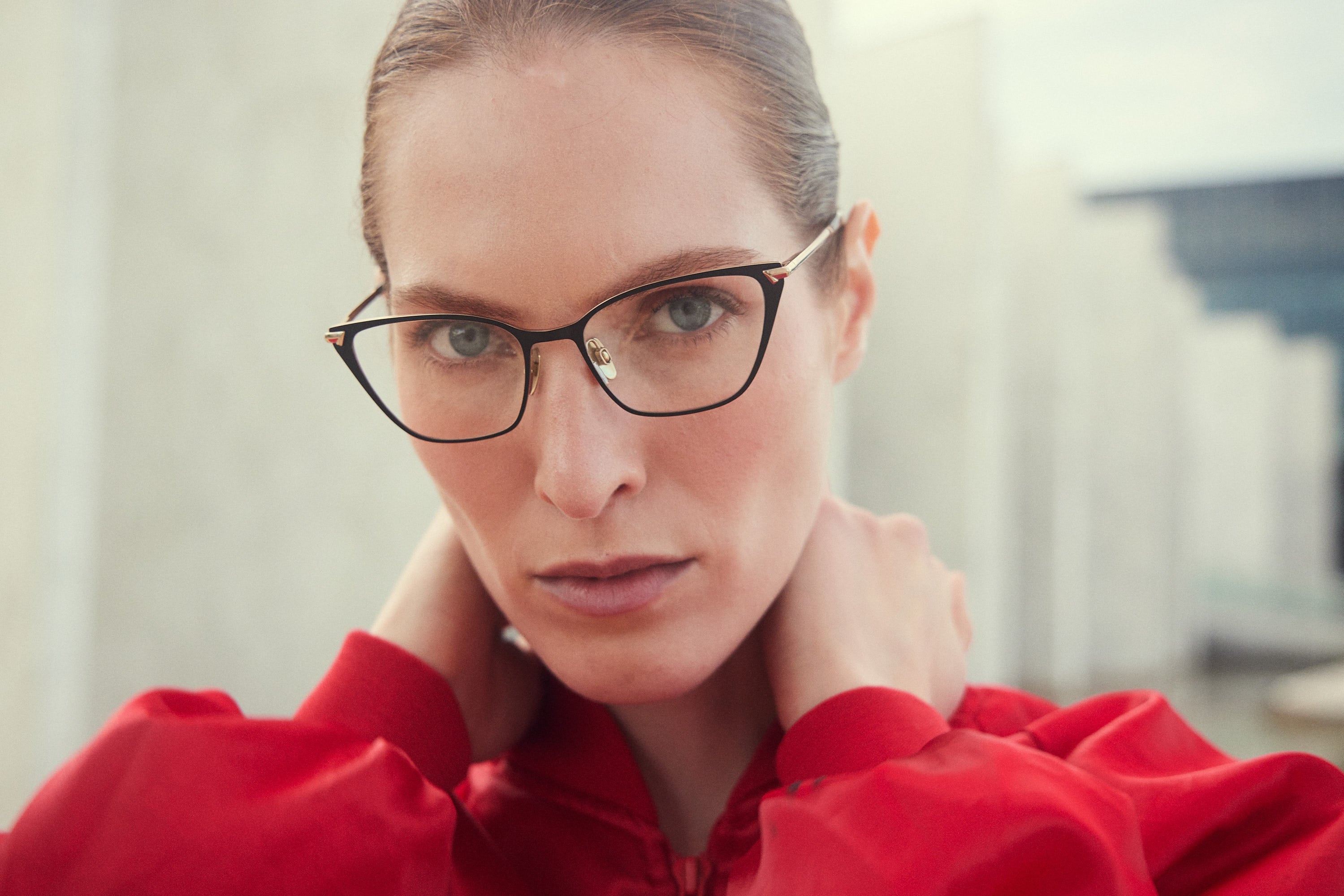 A close-up portrait of a woman wearing cat-eye titanium eyeglasses and a bright red top, with her hands behind her neck, set against a softly blurred light gray background.