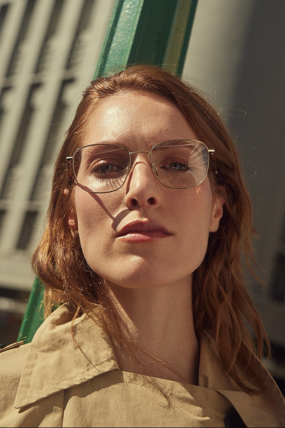 A close-up portrait of a woman with red hair wearing gold titanium eyeglasses and a beige trench coat, photographed outdoors with strong sunlight and shadows.