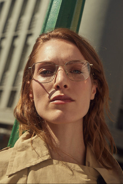 A close-up portrait of a woman with red hair wearing gold titanium eyeglasses and a beige trench coat, photographed outdoors with strong sunlight and shadows.