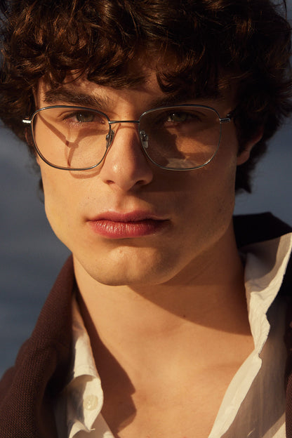 A close-up portrait of a young man with curly brown hair wearing silver square navigator titanium eyeglasses, a white collared shirt, and a dark brown sweater, set against a softly blurred outdoor background.