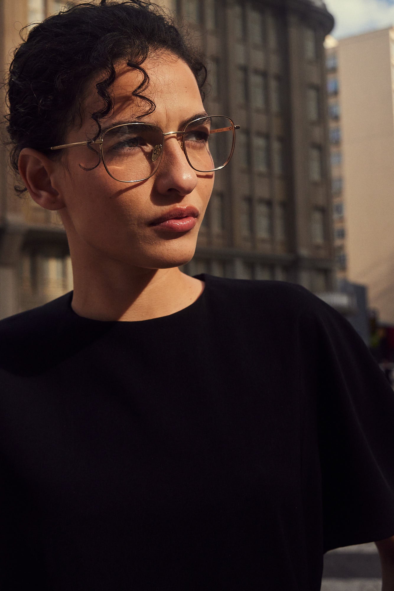 A woman outdoors wearing silver titanium eyeglasses with nosepads and a black crew-neck t-shirt, with a blurred city building in the background.