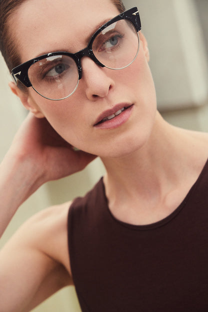 A close-up portrait of a woman wearing black marble cat-eye eyeglasses with silver metal wire rims, paired with a dark brown sleeveless top.
