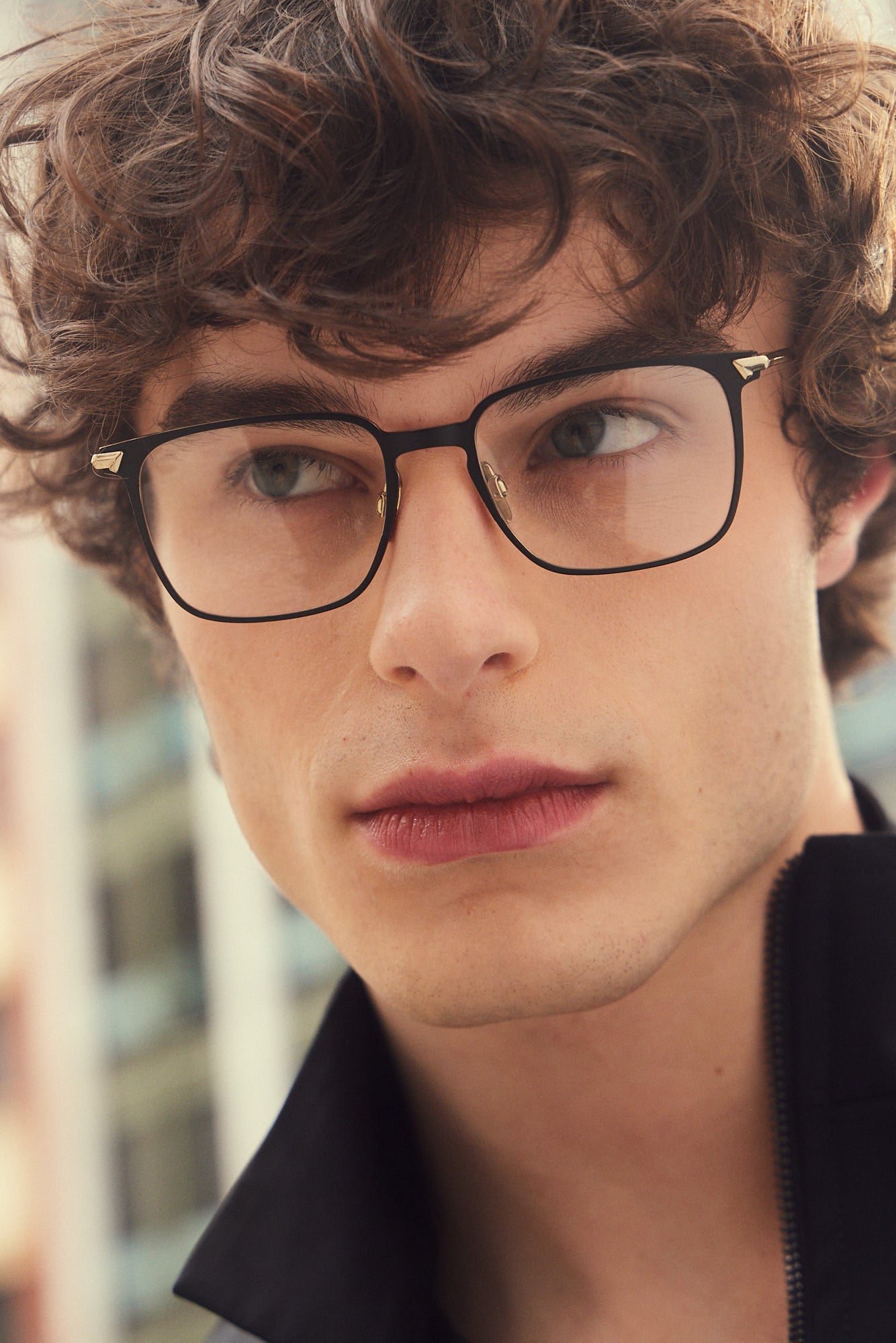 A close-up portrait of a young man with curly brown hair wearing black titanium rectangular eyeglasses with gold metal deatils on the hinges, set against a softly blurred outdoor background.