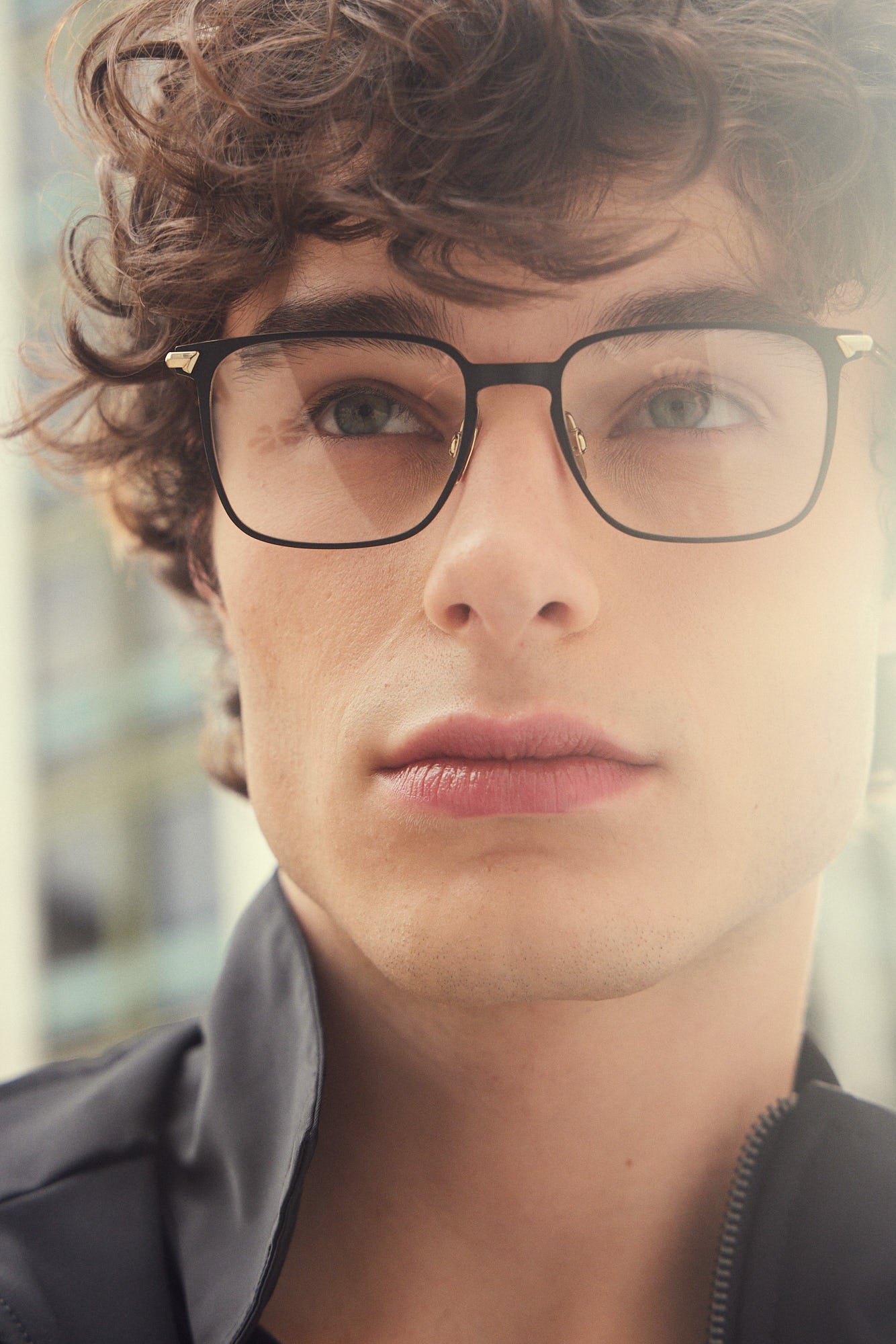A close-up portrait of a young man with curly brown hair wearing black rectangular titanium eyeglasses, photographed in soft natural light.