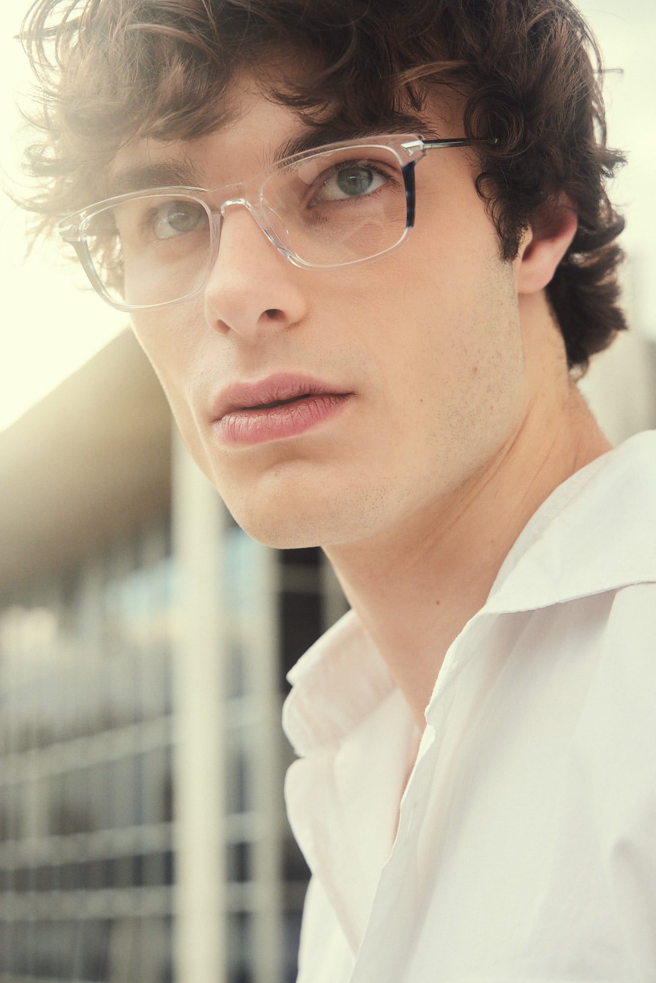 A close-up portrait of a young man with curly brown hair wearing clear-framed eyeglasses with silver titanium temples, wearing a white collared shirt, photographed outdoors with a softly blurred background.