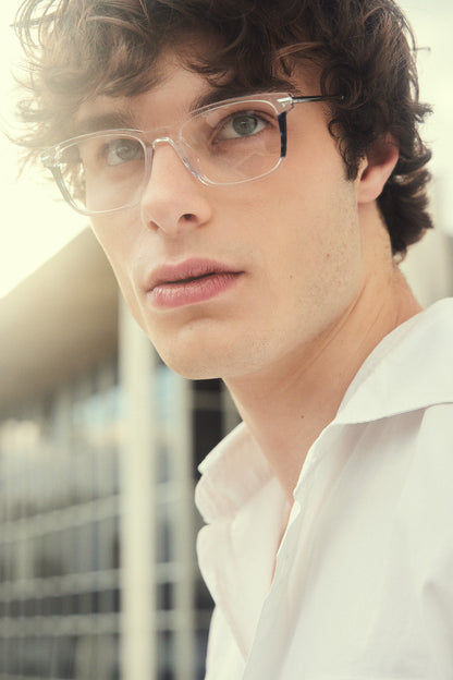 A close-up portrait of a young man with curly brown hair wearing clear-framed eyeglasses with silver titanium temples, wearing a white collared shirt, photographed outdoors with a softly blurred background.