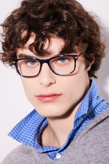 A close-up portrait of a young man with curly brown hair wearing navy and havana rectangular eyeglasses, a blue and white checkered shirt, and a gray sweater against a plain light background.