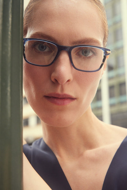 A close-up portrait of a woman wearing dark blue rectangular eyeglasses with havana accents, with a blurred outdoor city background.