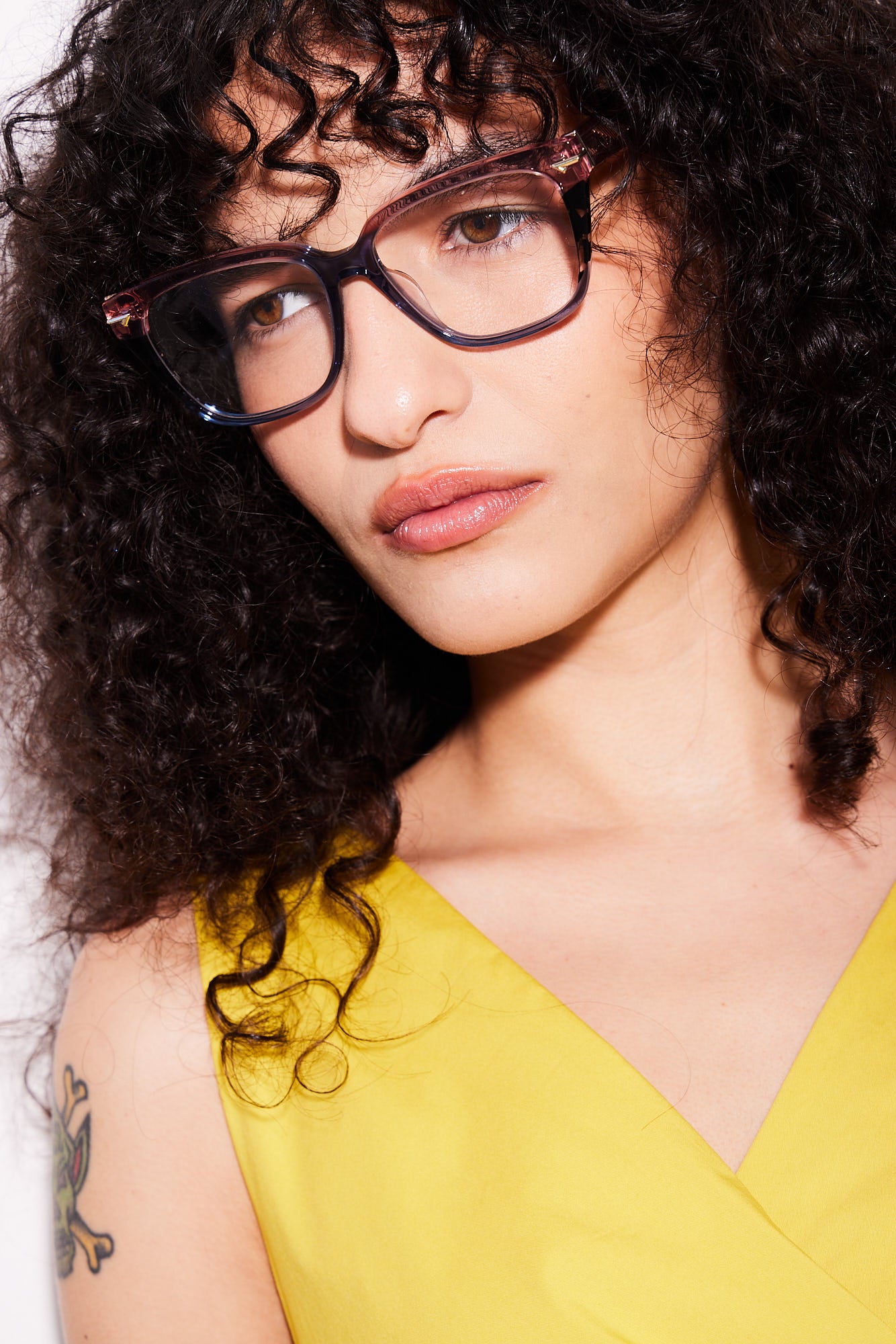 A close-up portrait of a woman with curly dark hair wearing rose blue gradient cateye eyeglasses and a bright yellow top.