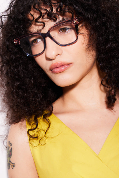 A close-up portrait of a woman with curly dark hair wearing rose blue gradient cateye eyeglasses and a bright yellow top.