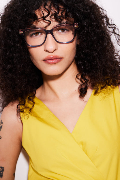 A close-up portrait of a woman with curly dark hair wearing trendy square eyeglasses with a subtle rose to blue gradient and a bright yellow top against a plain light background.