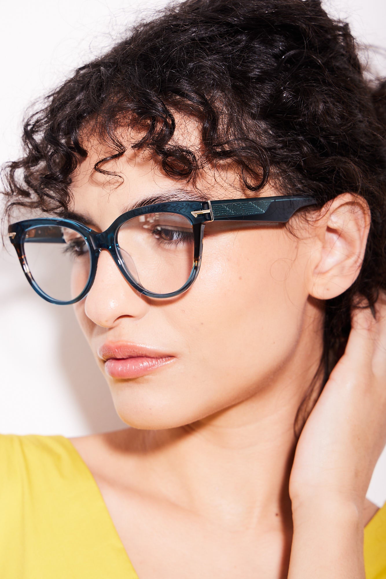 A close-up of a woman with curly dark hair wearing, oversized cat-eye teal acetate eyeglasses with  tortoiseshell pattern accents, paired with a bright yellow top against a plain white background.