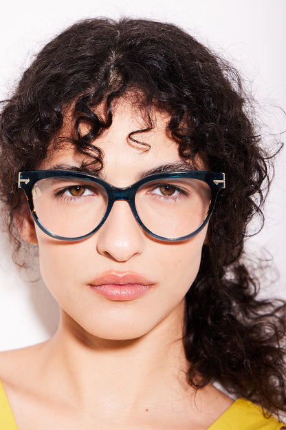 A close-up portrait of a model with curly dark hair wearing acetate teal cat-eye eyeglasses with silver metal hinges, set against a plain white background.