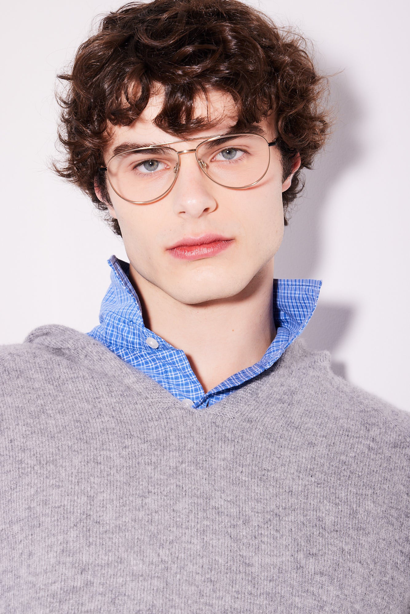 A close-up portrait of a young man with curly brown hair wearing aviator, titanium-framed eyeglasses, a blue checkered button-down shirt, and a gray knit sweater against a plain white background.