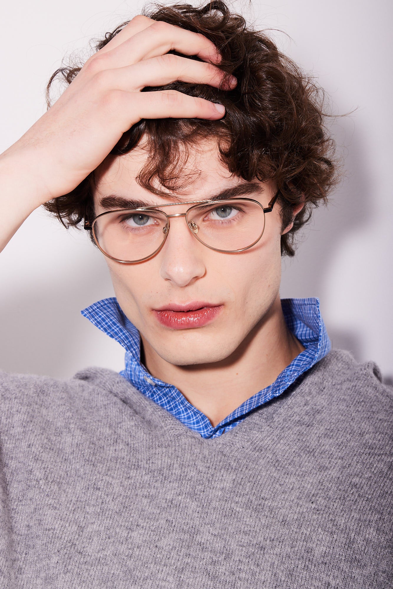 A close-up portrait of a young man with curly brown hair wearing gold aviator titanium eyeglasses, a gray knit sweater, and a blue checkered shirt collar, against a plain light background.
