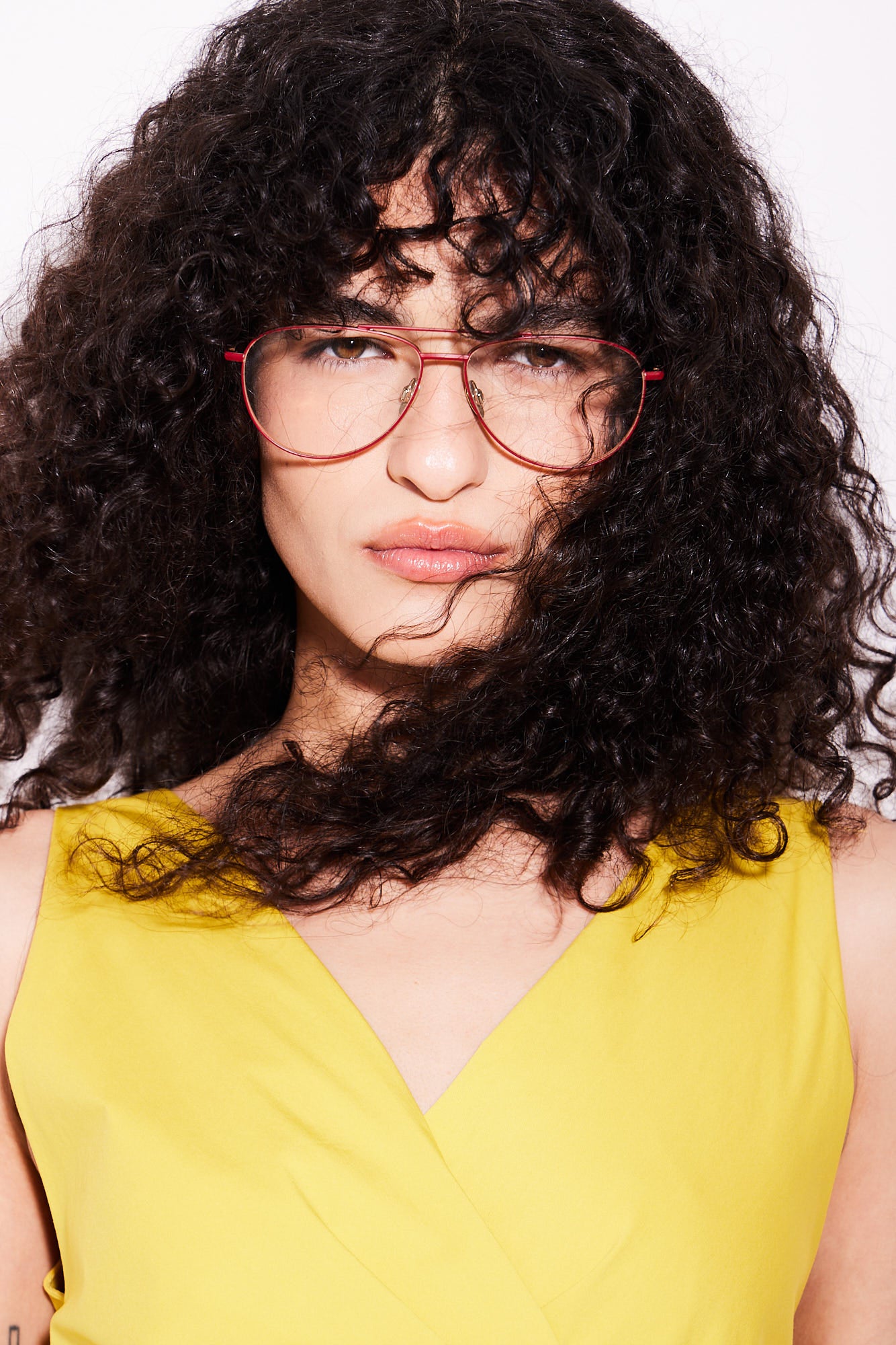A close-up portrait of a person with voluminous dark curly hair wearing red-framed aviator titanium eyeglasses and a bright yellow sleeveless top against a plain white background.