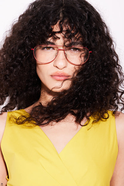 A close-up portrait of a person with voluminous dark curly hair wearing red-framed aviator titanium eyeglasses and a bright yellow sleeveless top against a plain white background.