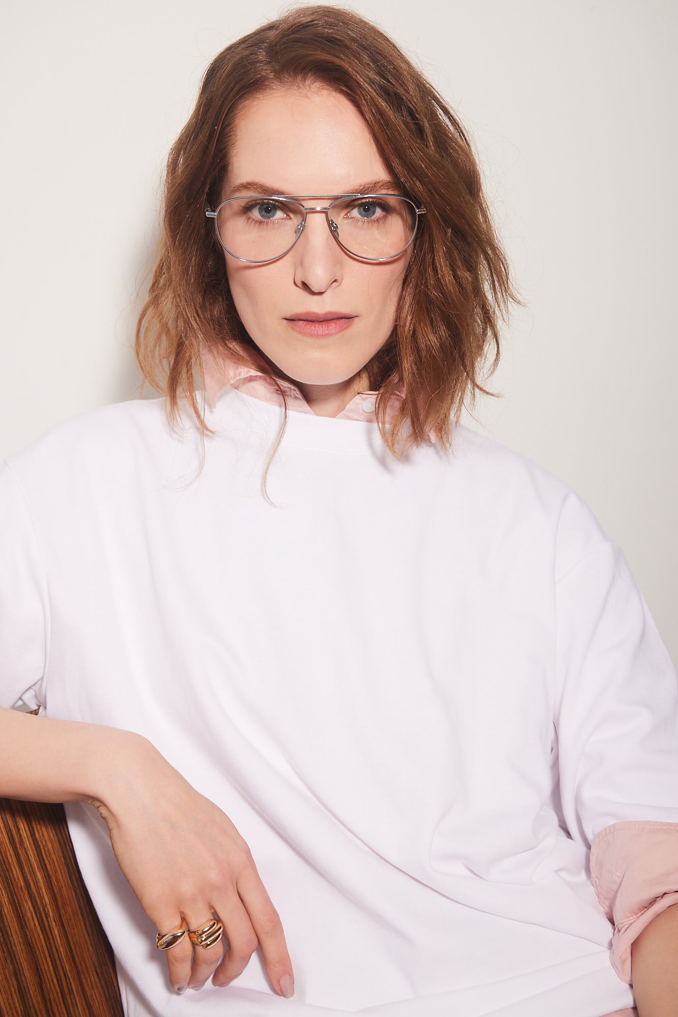 A woman with shoulder-length brown hair and quality aviator titanium-framed eyeglasses, wearing a white oversized sweatshirt with a pink inner collar, leaning against a wooden chair against a light beige background.