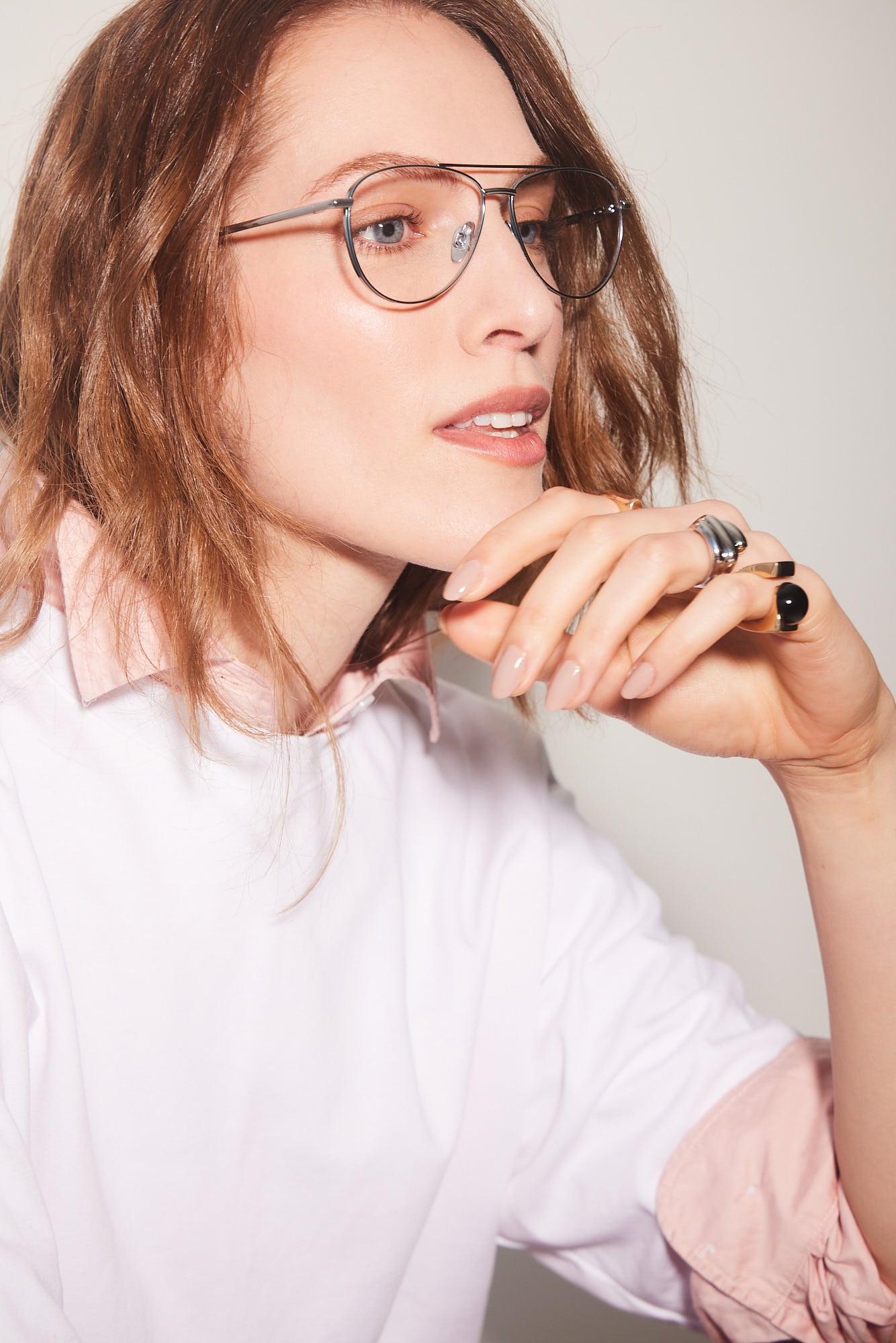 A close-up portrait of a woman with red hair wearing aviator titanium-framed eyeglasses, a white top with a light pink collar, and silver rings on her fingers, set against a plain light gray background.