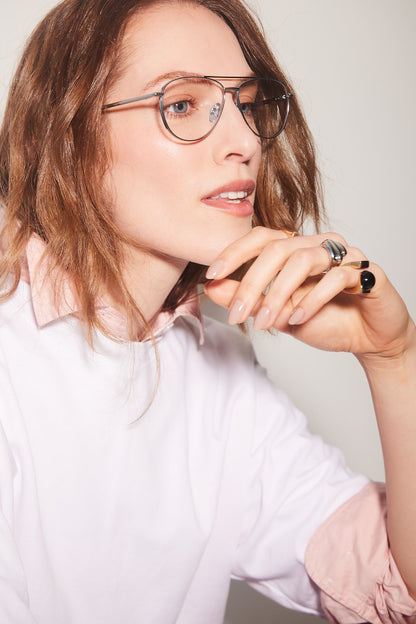 A close-up portrait of a woman with red hair wearing aviator titanium-framed eyeglasses, a white top with a light pink collar, and silver rings on her fingers, set against a plain light gray background.