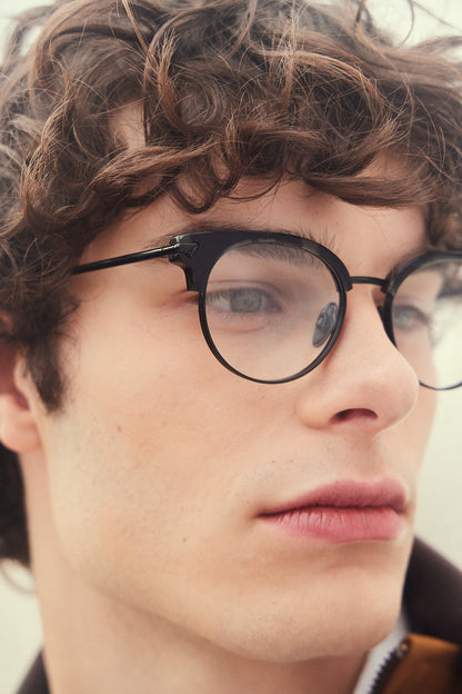 A close-up portrait of a young man with curly brown hair wearing black round clubmaster eyeglasses with titanium rims, temples and nosepads, against a soft light gray background.