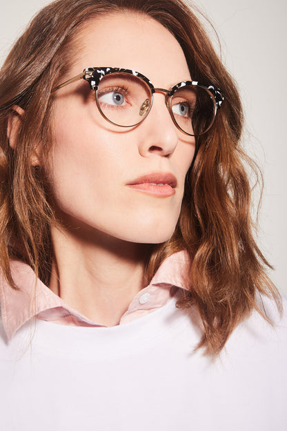 A close-up of a woman with wavy brown hair wearing round clubmaster eyeglasses with a black and white patterned top frame and gold titanium rims, paired with a light pink collared shirt against a light gray background.