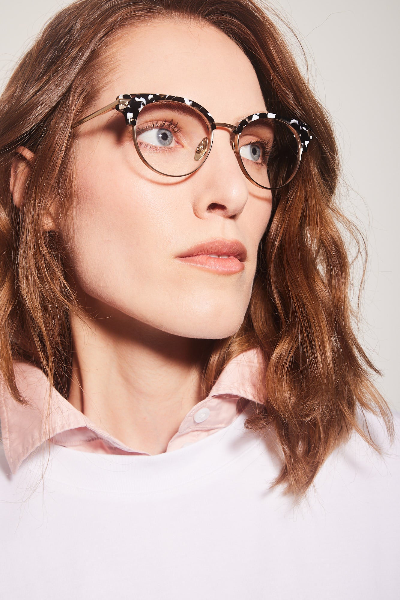A close-up of a woman with wavy brown hair wearing round clubmaster eyeglasses with a black and white patterned top frame and gold titanium rims, paired with a light pink collared shirt against a light gray background.