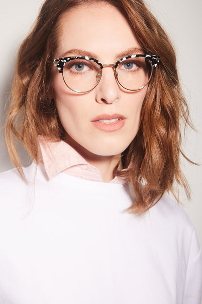 A close-up portrait of a woman with wavy light brown hair wearing round clubmaster eyeglasses with a black and white marble acetate and gold titanium rims, paired with a white top and a light pink collared shirt underneath.