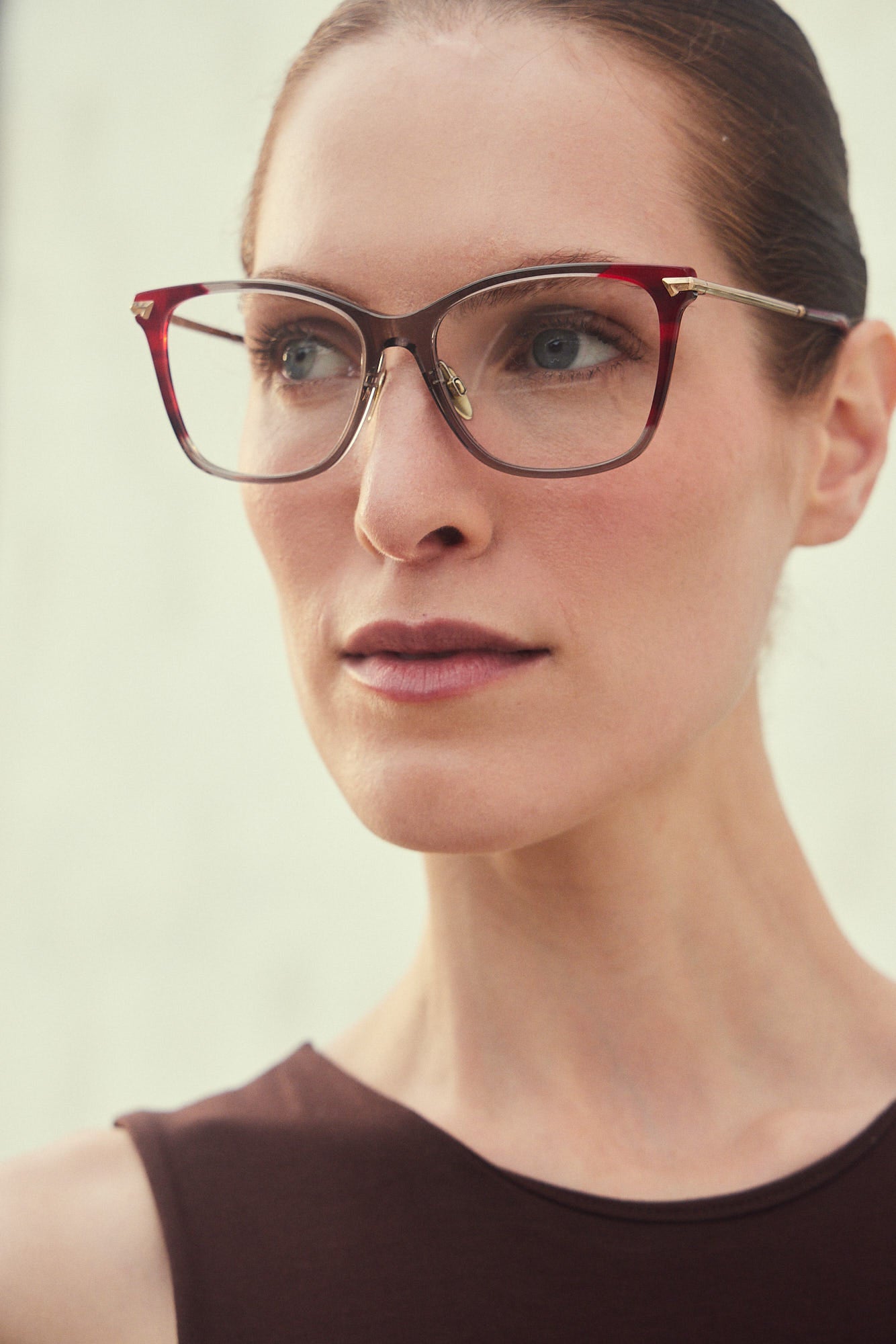 A close-up portrait of a woman wearing butterfly eyeglasses with a smoky gray  and red acetate frame with gold titanium temples, set against a soft, light beige background.