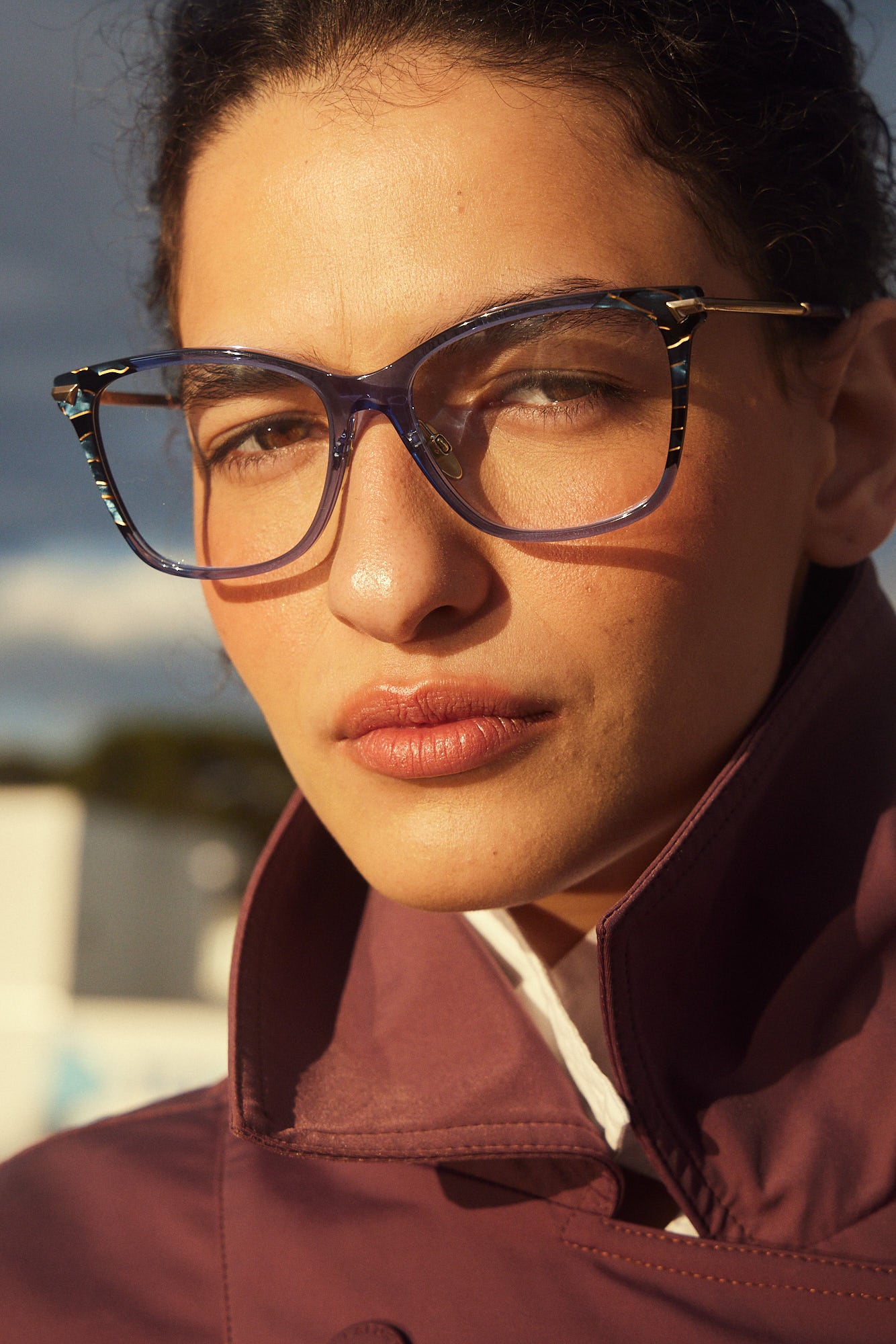 A close-up portrait of a woman wearing butterfly eyeglasses with a translucent dark blue acetate frame and gold titanium temples, set against a warm outdoor background.