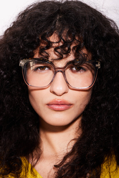 A close-up portrait of a woman with voluminous dark curly hair wearing oversized square acetate eyeglasses.