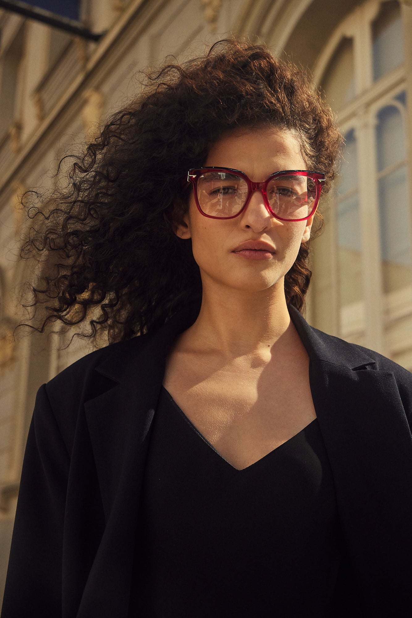 A woman with voluminous curly dark hair wearing pink-framed eyeglasses, a black top, and a black blazer, photographed outdoors against a sunlit building facade.
