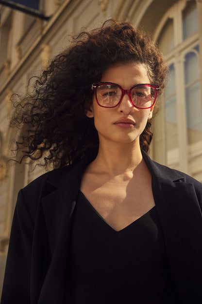 A woman with voluminous curly dark hair wearing pink-framed eyeglasses, a black top, and a black blazer, photographed outdoors against a sunlit building facade.