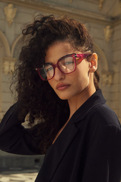 A woman with curly dark hair wearing an over sized square fuchsia acetate eyeglasses and a black blazer, standing outdoors in front of a stone building.