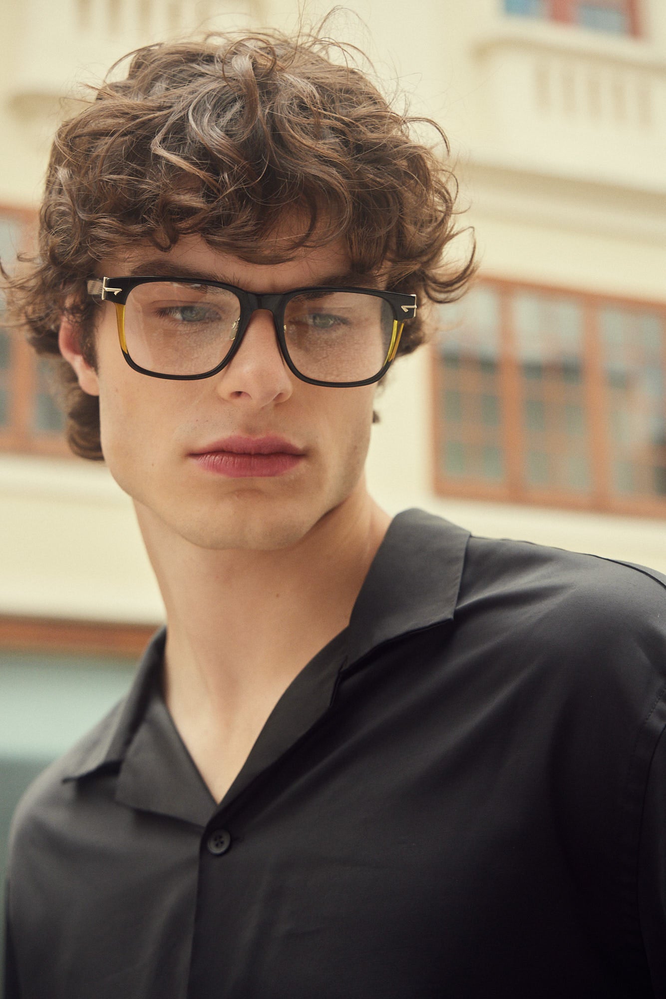 A close-up portrait of a young man with curly brown hair wearing black oversized square eyeglasses with sculpted rivets and a black button-up shirt, set against a softly blurred beige building background.