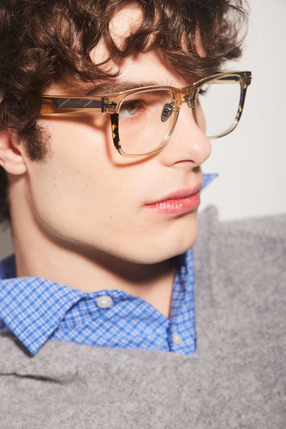 A close-up side profile of a man with curly brown hair wearing, oversized square khaki eyeglasses with titanium nosepads and tortoiseshell accents, styled with a blue checkered shirt and a gray sweater.