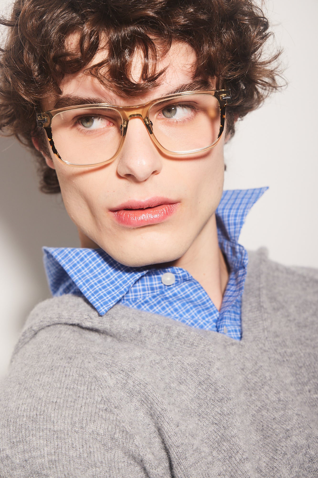 A close-up portrait of a person with curly brown hair wearing, oversized square khaki acetate eyeglasses with tortoiseshell accents, a blue checkered button-up shirt, and a gray knit sweater against a plain light background.