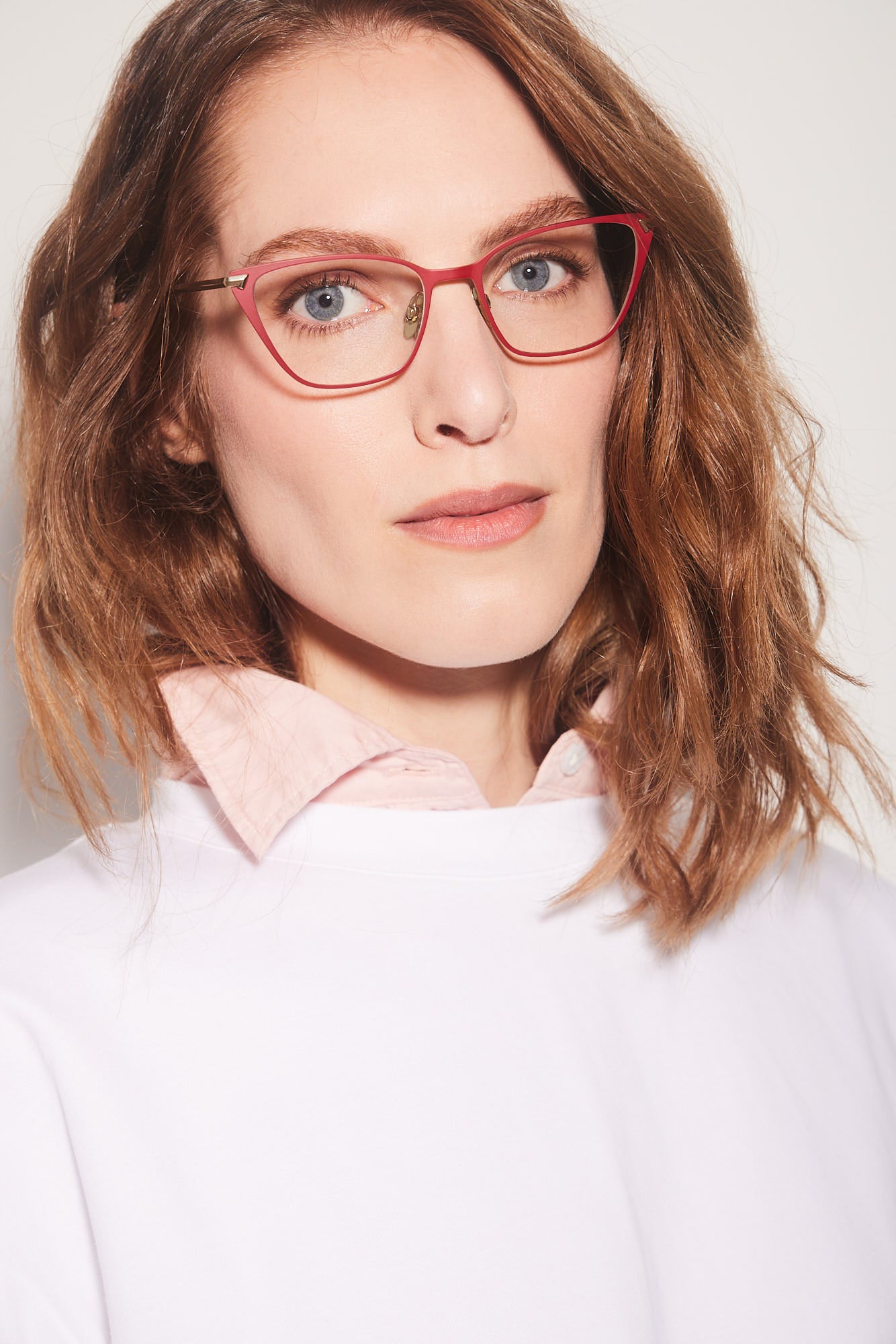 A close-up portrait of a woman with wavy light brown hair wearing red titanium cat-eye eyeglasses and a white top over a light pink collared shirt, set against a plain light gray background.