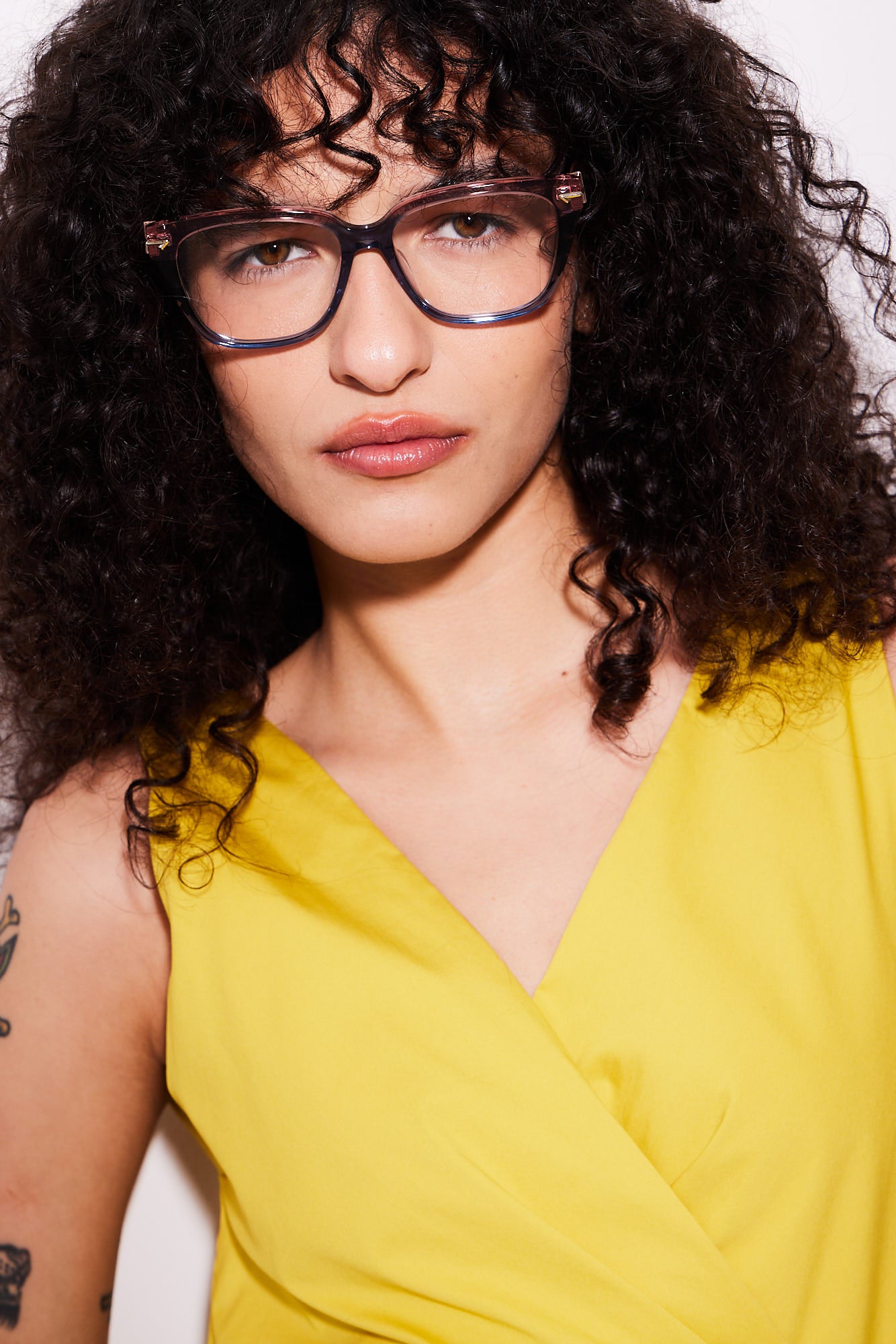 A close-up portrait of a woman with curly dark hair wearing oversized square acetate eyeglasses in a rose to blue gradient color and a bright yellow top against a plain light background.