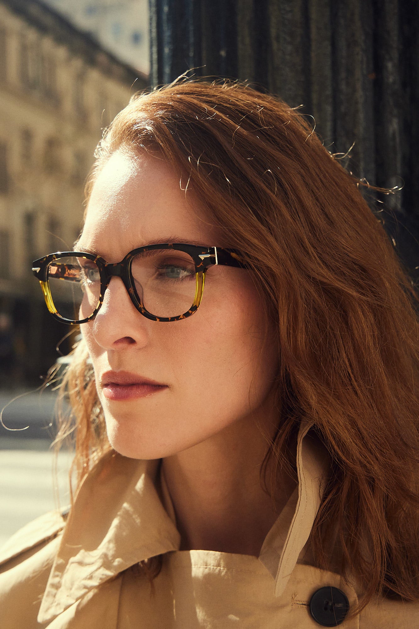 A close-up portrait of a woman with red hair wearing dark tortoiseshell acetate eyeglasses and a beige trench coat, set against an outdoor urban background.