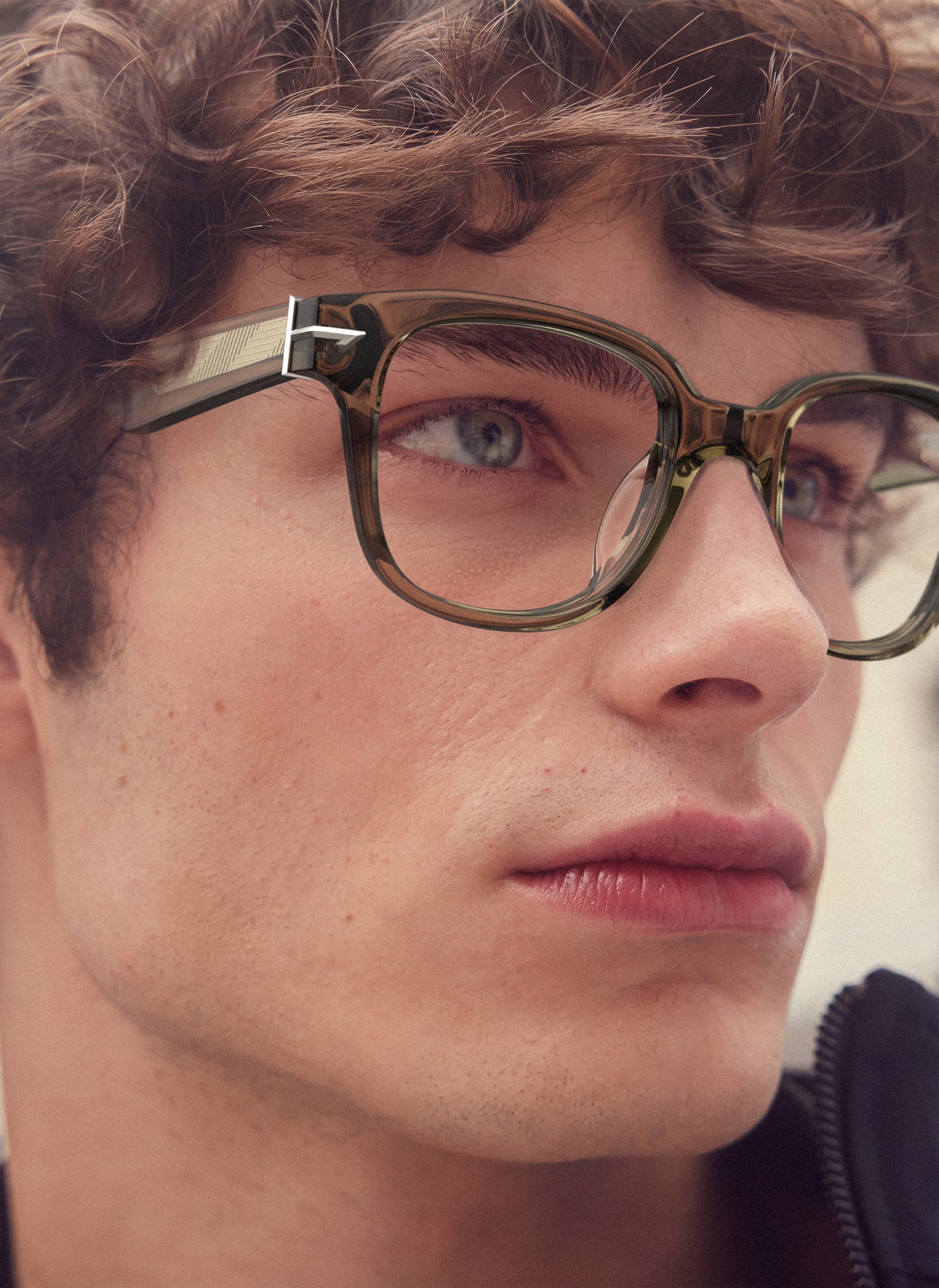 A close-up portrait of a young man with curly brown hair wearing, translucent olive green acetate eyeglass frames with a silver metal hinge detail.